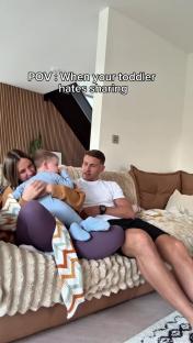 A family sits on a couch; a toddler is held by his mother while his father looks on. Text overlay reads: POV: When your toddler hates sharing.