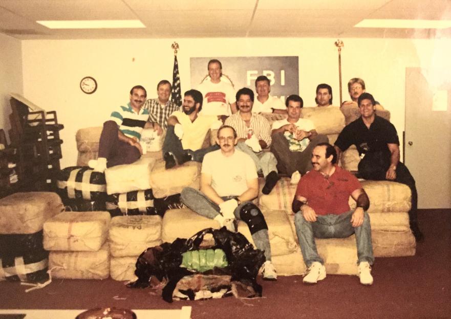 Men posing with bales of illegal narcotics in front of an FBI logo.