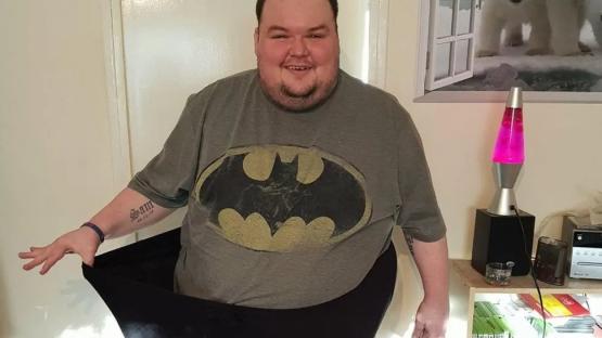 Man in Batman shirt holding up large pants to show weight loss.