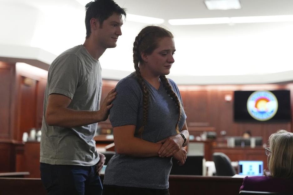 A man stands behind a woman with a hand on her shoulder in a courtroom.