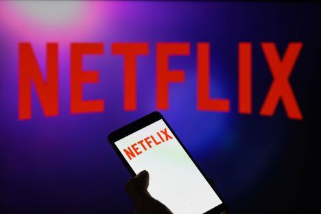 Photo illustration of a smartphone displaying the Netflix logo, with a larger Netflix logo visible on a screen in the background.