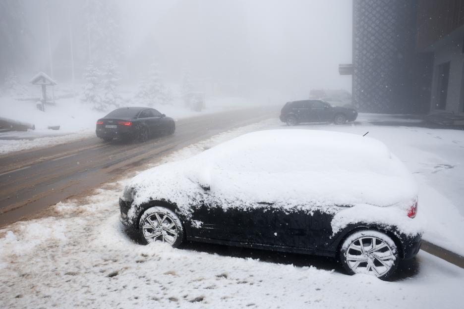 A snow-covered car parked next to a slushy road where another car drives through a snowfall.