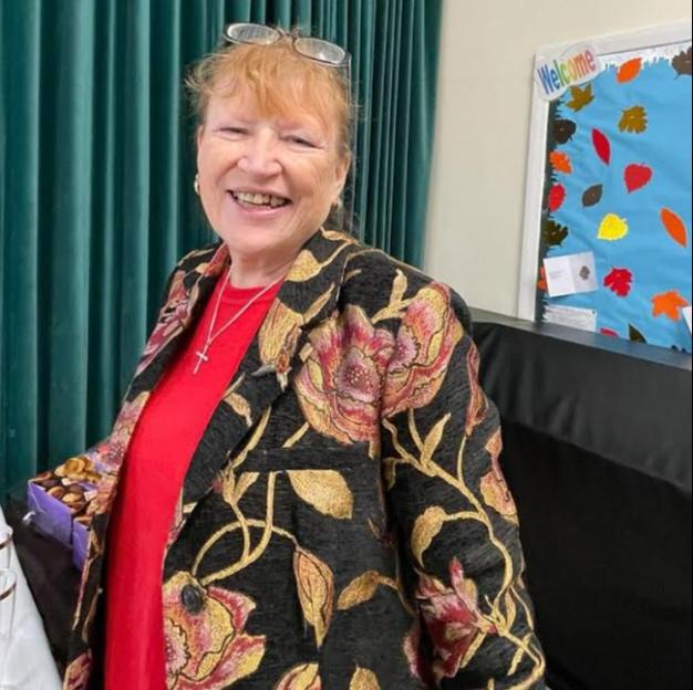 A smiling woman with glasses on her head wears a black jacket with a gold and red floral pattern over a red shirt, with a silver cross necklace.