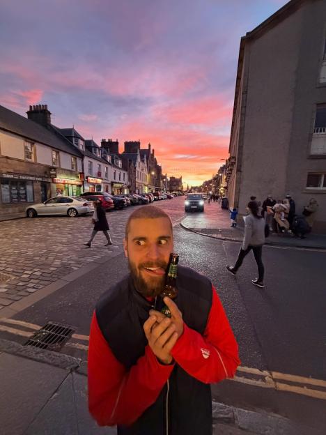 Man holding a bottle in a funny pose on a street with a pink and orange sunset.
