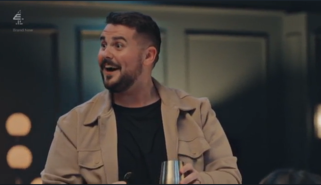 A man with a beard, wearing a beige jacket and black shirt, holds a silver cup and looks up to the right with his mouth open in an excited expression.