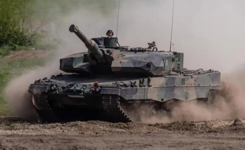 A Leopard 2 tank with a visible gunner, kicking up dust as it moves.