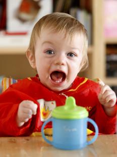 A baby wearing a red bib opens its mouth wide while sitting at a high chair with a sippy cup in front of it.