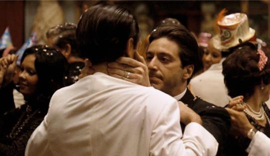 Scene from The Godfather Part II showing Al Pacino embracing another man at a party.