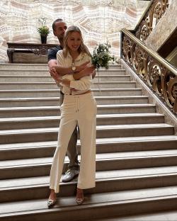 Couple embracing on a staircase, the woman wearing a white tracksuit and holding a bouquet.