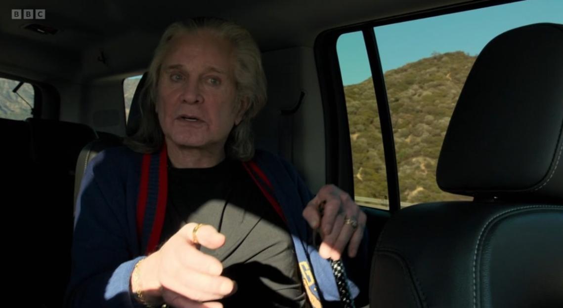 Ozzy Osbourne sits in the backseat of a car, speaking to someone off-camera.