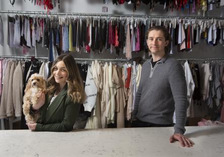 a woman holding a dog stands next to a man in front of clothes racks