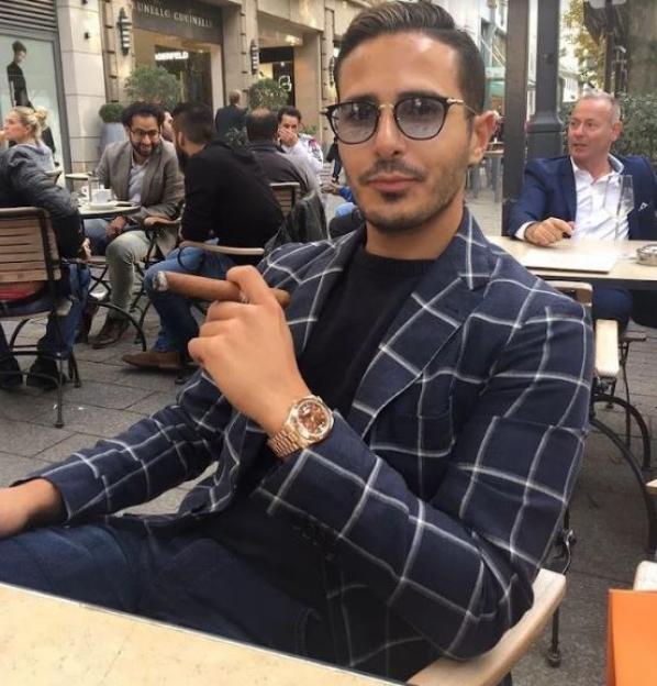 Simon Leviev, the "Tinder Swindler," in a checkered blazer, holding a cigar and wearing a gold watch.