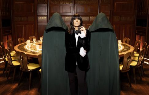 Claudia Winkleman on the set of The Traitors, flanked by two figures in hooded robes.