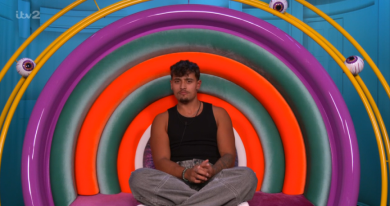 Big Brother housemate in a black tank top and grey jeans sitting on a multi-colored chair.