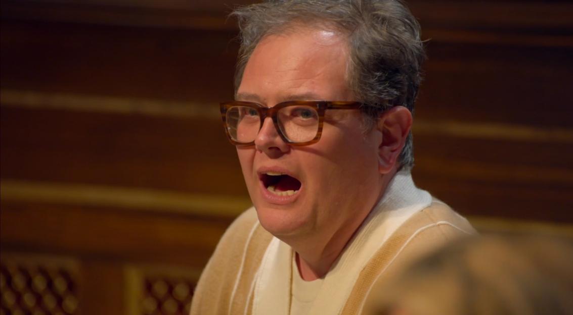 Alan Carr from The Traitors with his mouth open, wearing a beige and cream collared sweater.