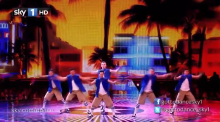 Six male dancers in blue jackets, tan pants, and white socks with black stripes on stage with a backdrop of a city street with palm trees.