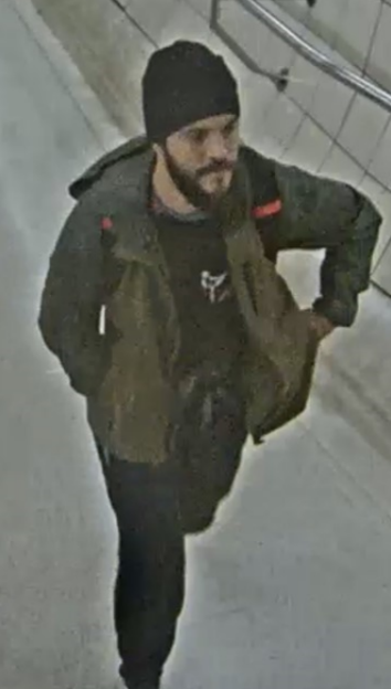 CCTV image of a man with a beard and dark beanie, wearing a dark jacket, walking down a tiled hallway.