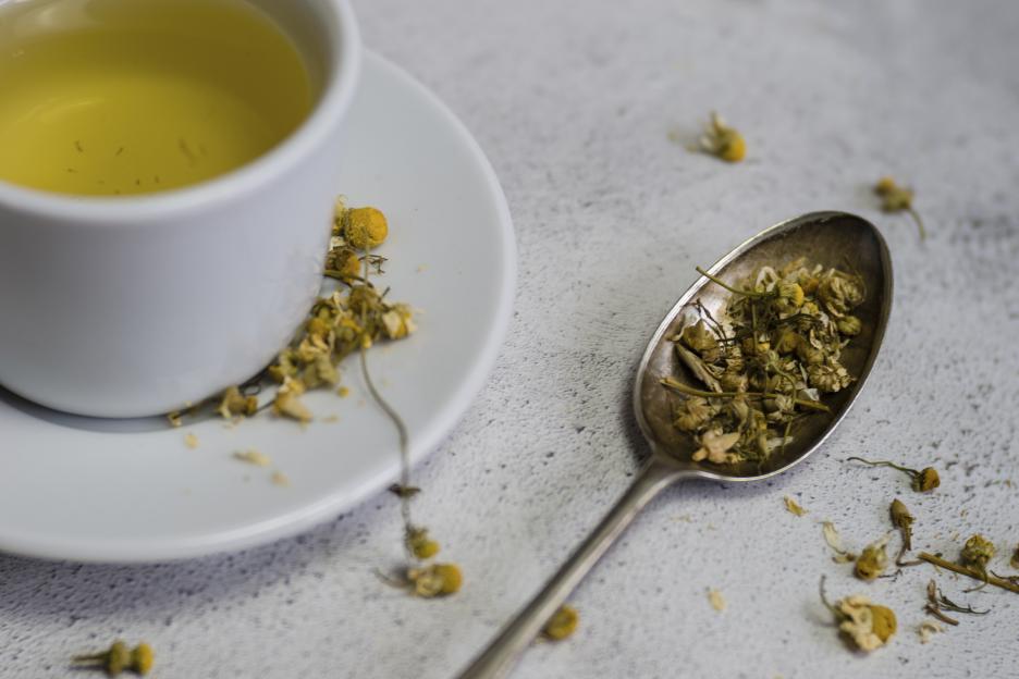 A high-angle view of a white cup of chamomile tea and a spoon with dried chamomile flowers.