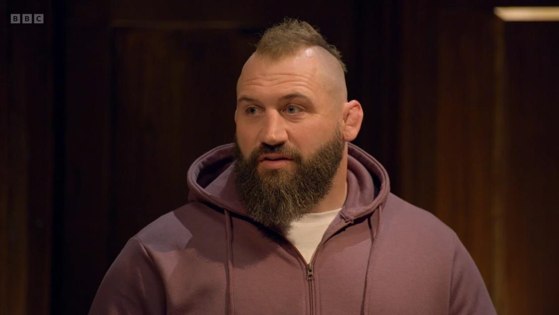 A man with a mohawk, beard, and purple hoodie looks to his left.