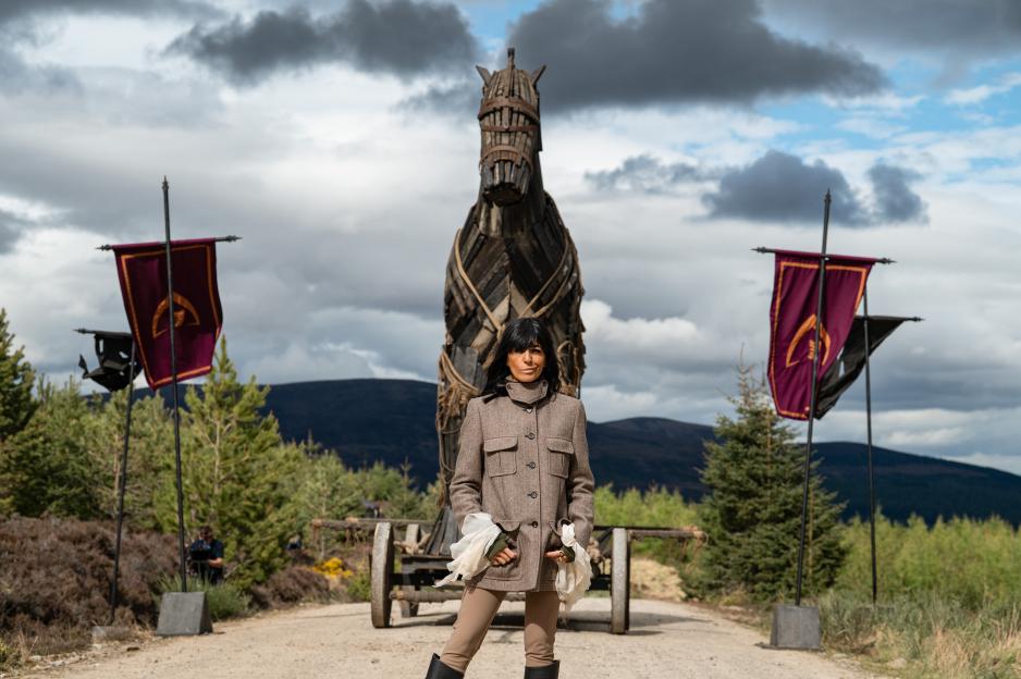 A presenter in a brown coat stands in front of a large wooden Trojan horse-like structure, flanked by two flags, with mountains and pine trees in the background.