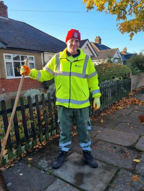 Wayne Broadhurst smiling, wearing a high-visibility jacket, a red beanie, and holding a broom.