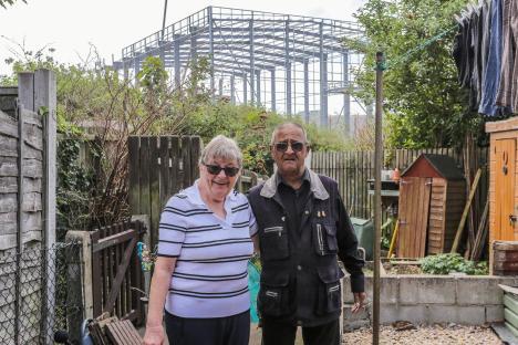 A couple stands in their garden with a large warehouse under construction visible in the background.