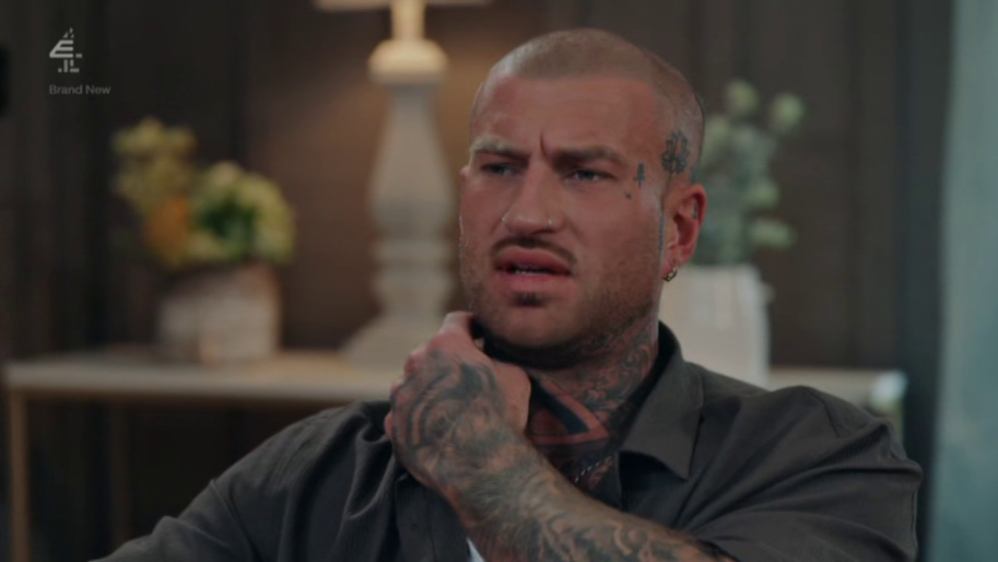 A man with face and neck tattoos, wearing a gray shirt, holds his throat with one hand.