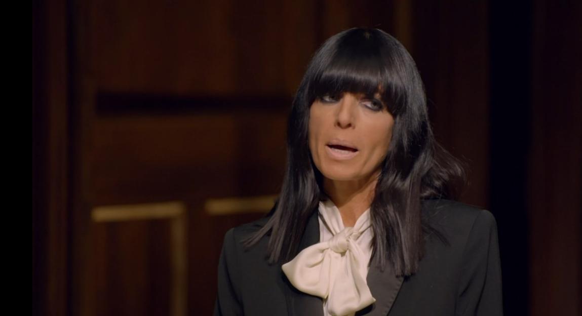 A woman with dark hair and heavy eye makeup, wearing a black suit jacket and a cream-colored bow-tie blouse, speaks.