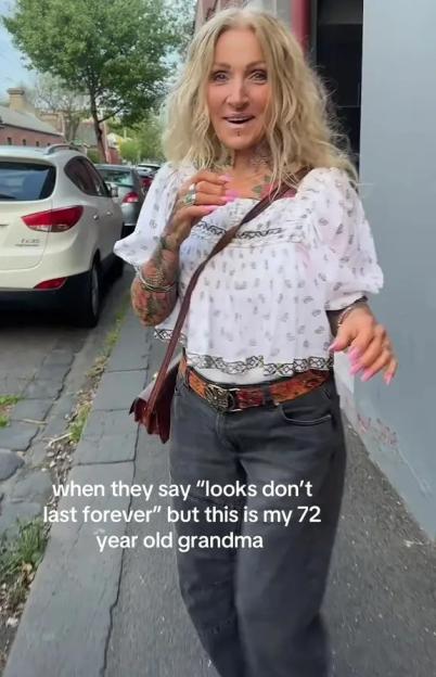 A 72-year-old woman with long blonde hair, tattoos, and piercings, wearing a white shirt and dark pants, walking on a sidewalk.