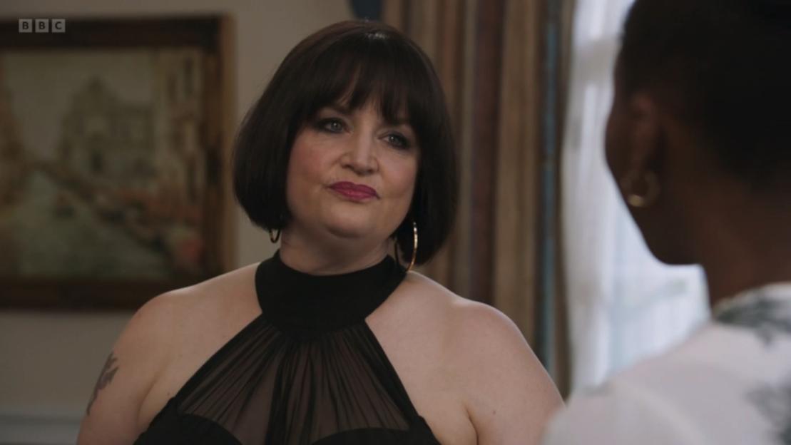 Nessa (Ruth Jones) from Gavin and Stacey in a black dress with an off-the-shoulder mesh top, looking towards someone.