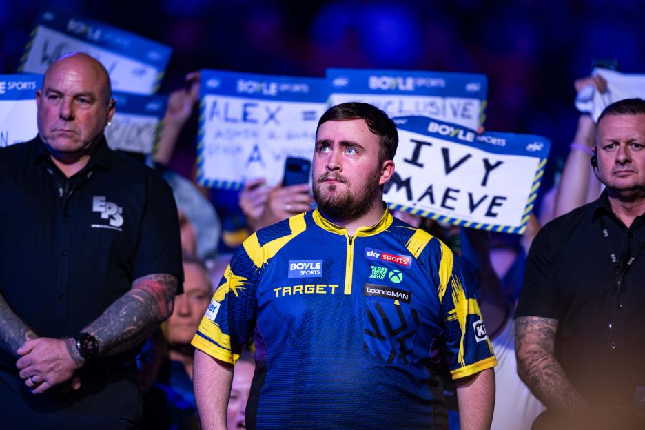 Luke Littler in a blue and yellow darts jersey, standing between two security guards.