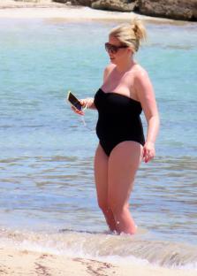 Woman in black swimsuit standing in shallow water, looking at her phone.
