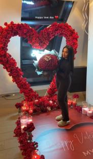 Woman standing in front of a heart-shaped rose arrangement holding a bouquet of roses that says