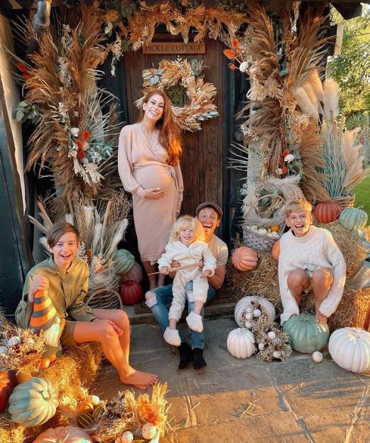 A pregnant woman, a man, and three children in fall decor with pumpkins and hay.