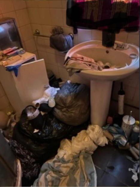 An extremely messy bathroom with a sink, toilet, and many overflowing trash bags and clothes on the floor.