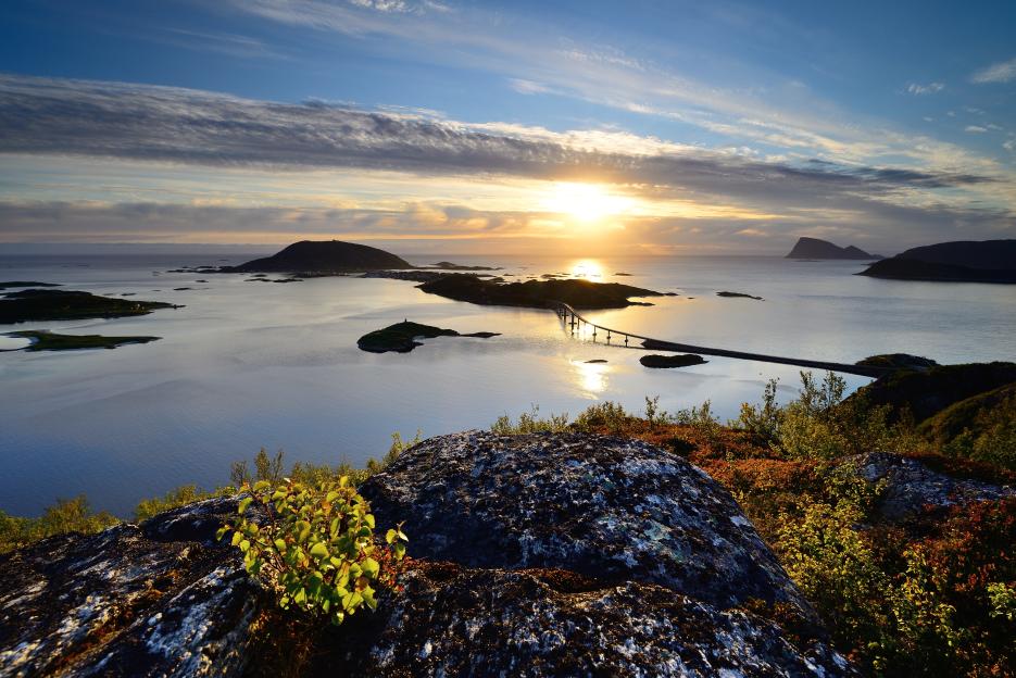 Sunset view of a bridge over water, with islands and mountains in the background.