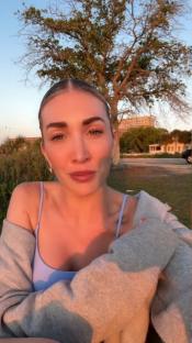 Woman in a light blue top and gray cardigan speaks to the camera outdoors at sunset.