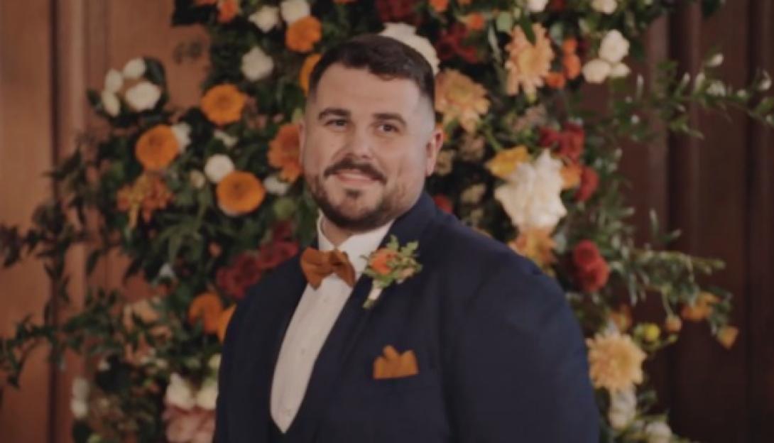 Groom in a navy suit with an orange bowtie and boutonniere, smiling in front of a floral arrangement.