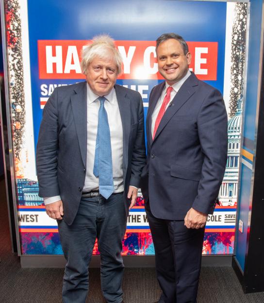 Boris Johnson and Harry Cole standing in front of a blue and red sign for "Harry Cole Saves the West" TV show.