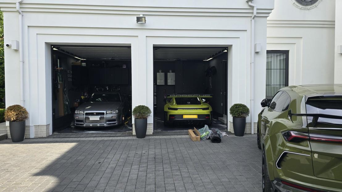Silver Nissan Skyline GT-R and a yellow Porsche 911 GT3 parked in a garage, with an olive green Lamborghini Urus parked outside.