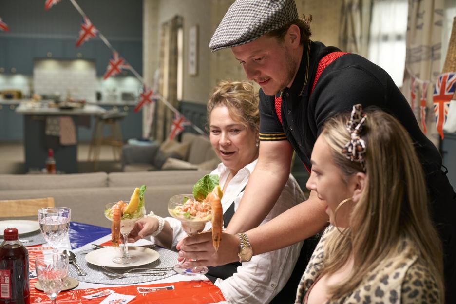 Charlie serves shrimp cocktails to Nicole and Isabelle at a table decorated with Union Jack flags.