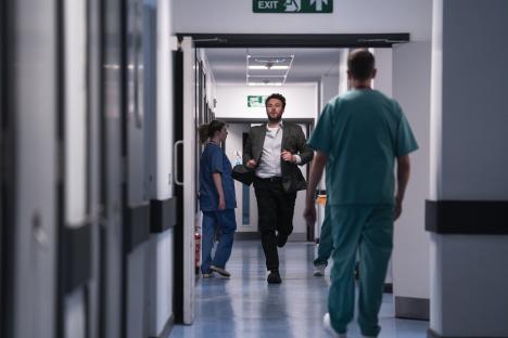 A man in a suit runs down a hospital corridor, followed by a nurse.