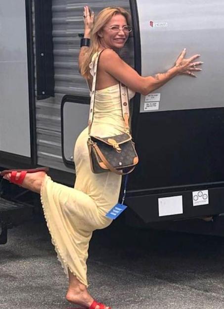 A woman in a long yellow dress, red sandals, and a shoulder bag smiles and leans against a grey trailer, one leg bent at the knee.