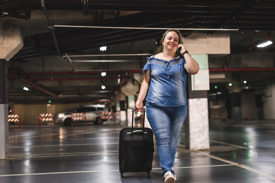 Woman in a parking garage talking on the phone and pulling a suitcase.