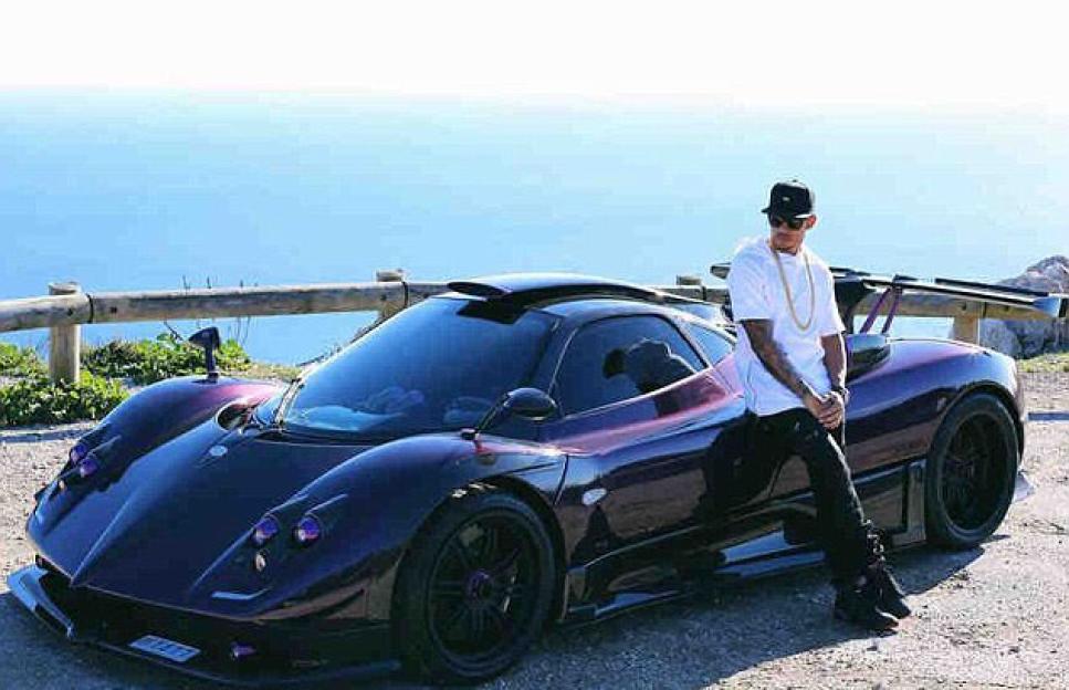 Lewis Hamilton posing next to a purple Pagani Zonda on a road overlooking the sea.