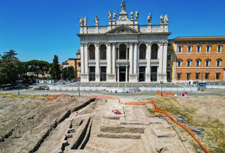 Excavated ruins of an ancient papal palace in Rome, in front of a basilica.
