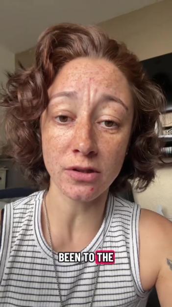 A woman with freckles and brown curly hair looks directly at the camera, wearing a striped tank top and a silver necklace.