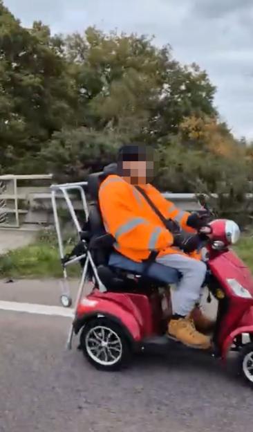 An elderly man riding a red mobility scooter on a busy motorway, wearing an orange high-visibility jacket.