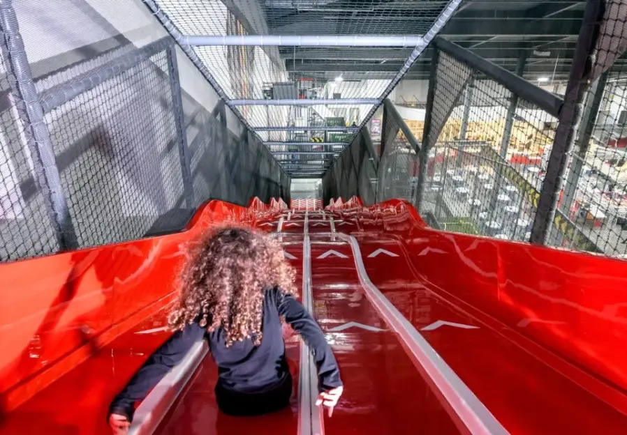 A child with curly hair sliding down a red multi-lane slide at an indoor play area.