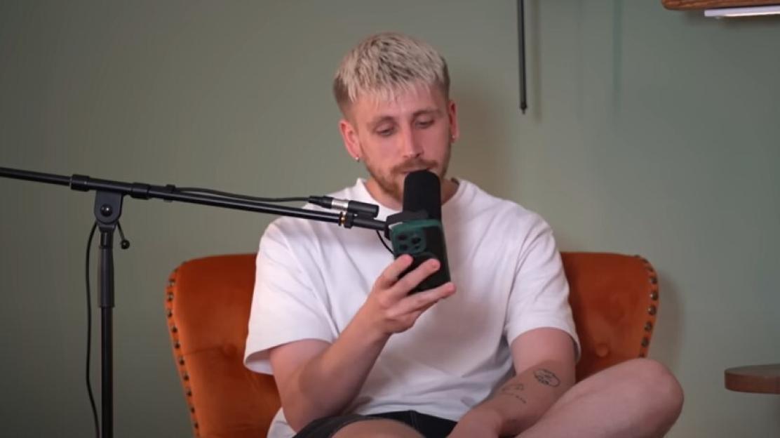 Man sitting in a chair looking at his phone while speaking into a microphone.